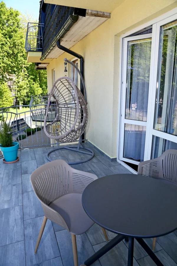 Balkon z wiszącym wiklinowym fotelem-kokonem, okrągłym stolikiem i krzesłem; drzewa i turkusowa donica z rośliną w tle.