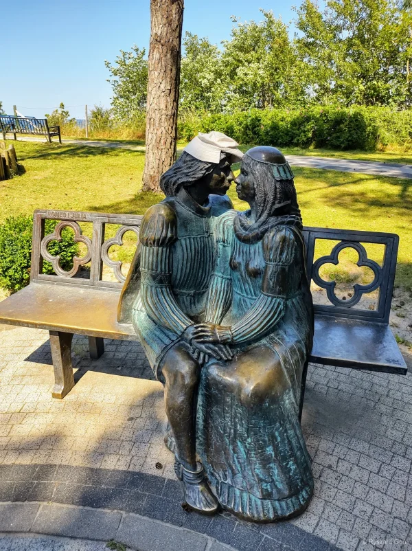 Brązowa rzeźba zakochanej pary na ławce z inskrypcją Romeo i Julia w parku w Rewalu, w tle morze.