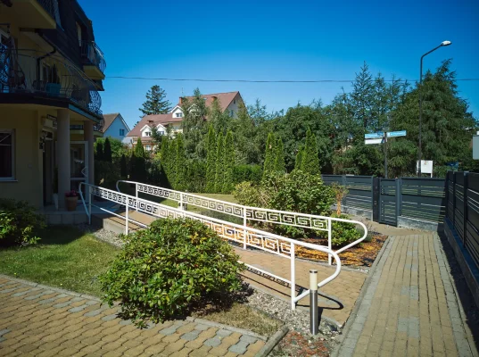 Wejście do pensjonatu z białą ażurową balustradą wzdłuż rampy, brukowaną alejką i zielonymi krzewami przed budynkiem.