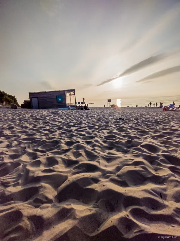 Plaża w Rewalu o zmierzchu: drewniany posterunek ratownika, ślady stóp na piasku i słońce odbijające się w Bałtyku.