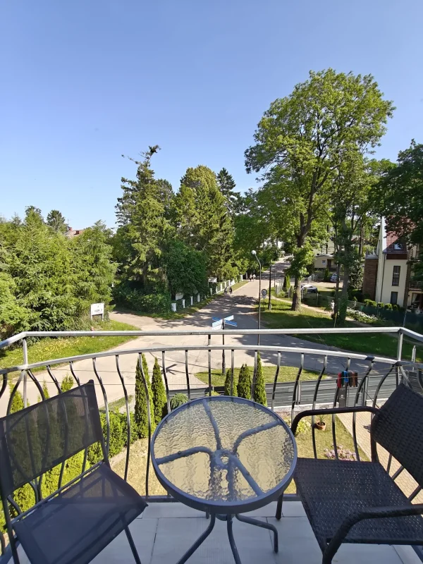 Widok z balkonu Pensjonatu Nadmorski w Rewalu na zadrzewioną ulicę; szklany stolik i wiklinowy fotel na balustradzie.
