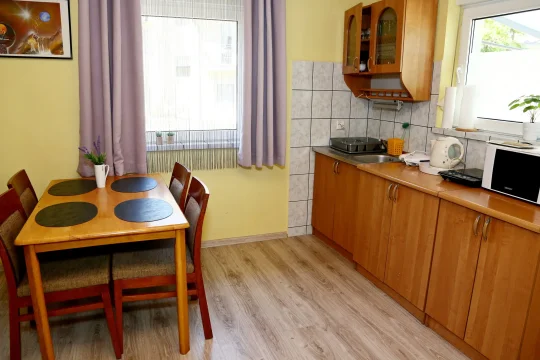 Drewniany stół z czterema krzesłami przy oknie oraz aneks kuchenny z szafkami, czajnikiem i mikrofalówką w salonie domku.