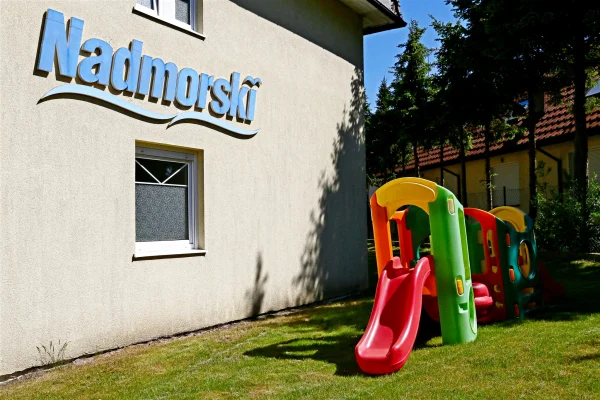 Kolorowa zjeżdżalnia i plastikowy plac zabaw dla dzieci na trawniku przy ścianie Pensjonatu Nadmorski w Rewalu.