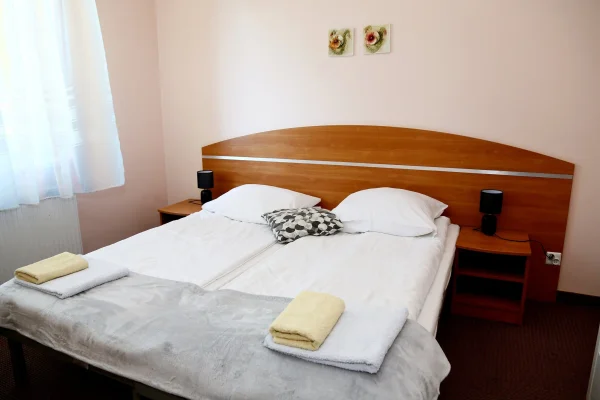 Podwójne łóżko z białą pościelą, szarą narzutą i złożonymi ręcznikami, drewniane wezgłowie ozdobione obrazami z kwiatami.